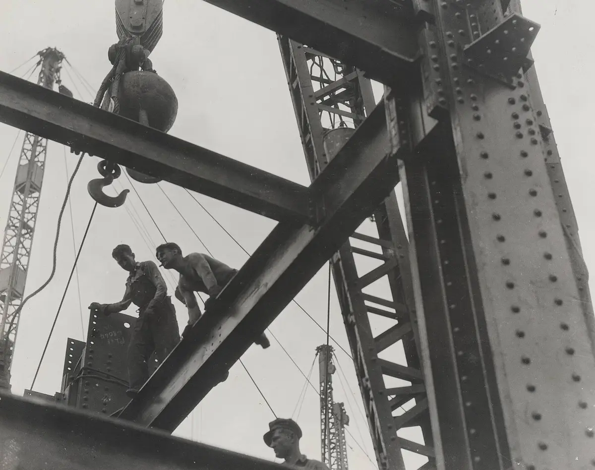 Ironworkers climb the steel frame of the Empire State Building beneath a derrick hook, 1930.