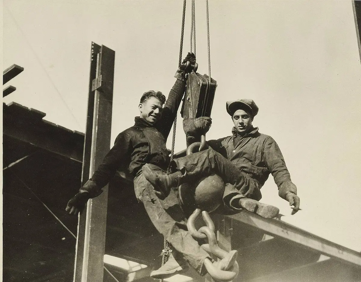 Two young structural workers ride a derrick hook on the rising frame of the Empire State Building.