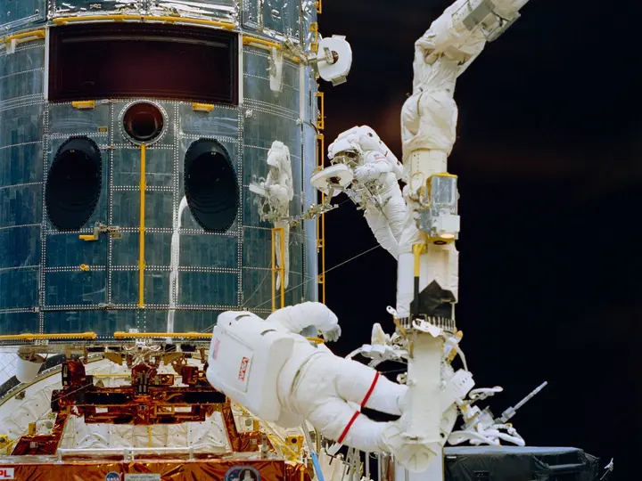 Jeffrey Hoffman, anchored to the end of Endeavour’s robot arm, prepares to install the new Wide Field and Planetary Camera 2 as Story Musgrave works at the open bay, 7 December 1993 (STS061-74-046).