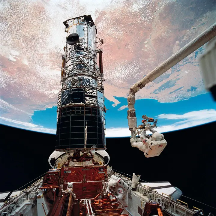 Story Musgrave rides the robot arm toward the top of Hubble on the fifth and final spacewalk, with Hubble standing upright in Endeavour’s payload bay above Earth, 9 December 1993 (STS061-98-050).