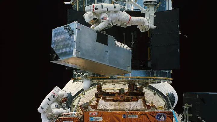 Kathryn Thornton lifts COSTAR, the corrective optics that gave Hubble’s instruments their sharp focus, while Tom Akers works below in Endeavour’s payload bay on the fourth spacewalk, 8 December 1993 (STS061-47-014).