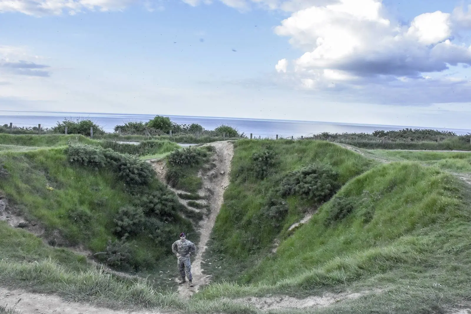 A paratrooper of the 173rd Airborne Brigade stands in a naval gunfire crater on Pointe du Hoc in 2018, giving scale to the ground the Rangers fought across.