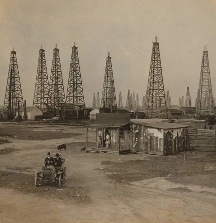 An early automobile rolls past oilfield shacks and derricks at Spindletop, about 1915